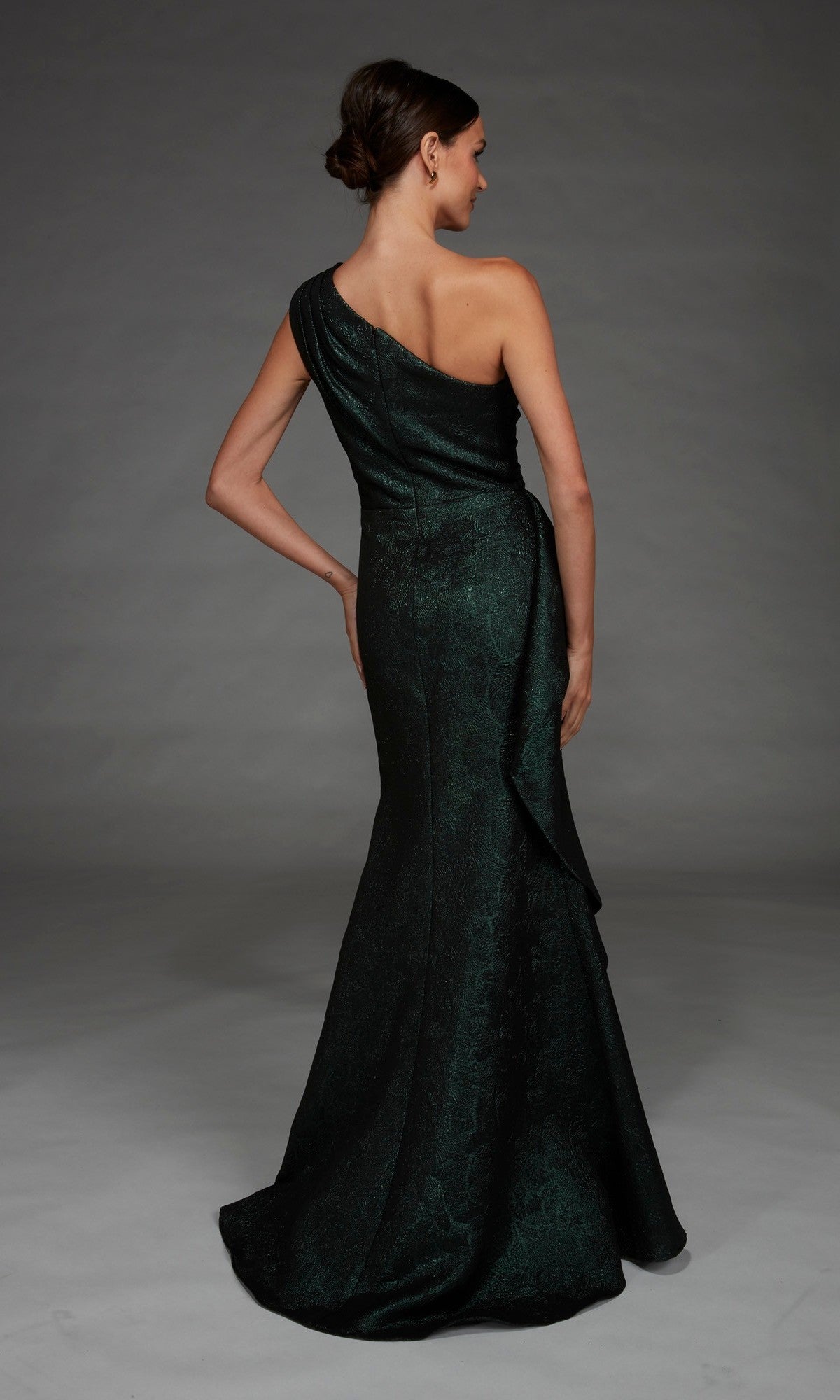 A woman stands with her back to the camera against a gray background, modeling the Alyce 27885 long formal dress in dark green, featuring a one-shoulder design.